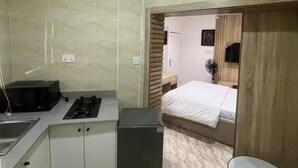 Cocina compacta con microondas, estufa y pileta. Se ve una cama con colchón blanco a través de una puerta abierta.
