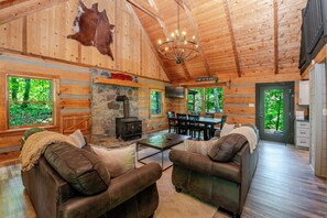 Ein Blockhaus-Interieur mit Kamin aus Stein, Ledersofas, einem Essbereich und großen Fenstern mit Blick auf den Wald.