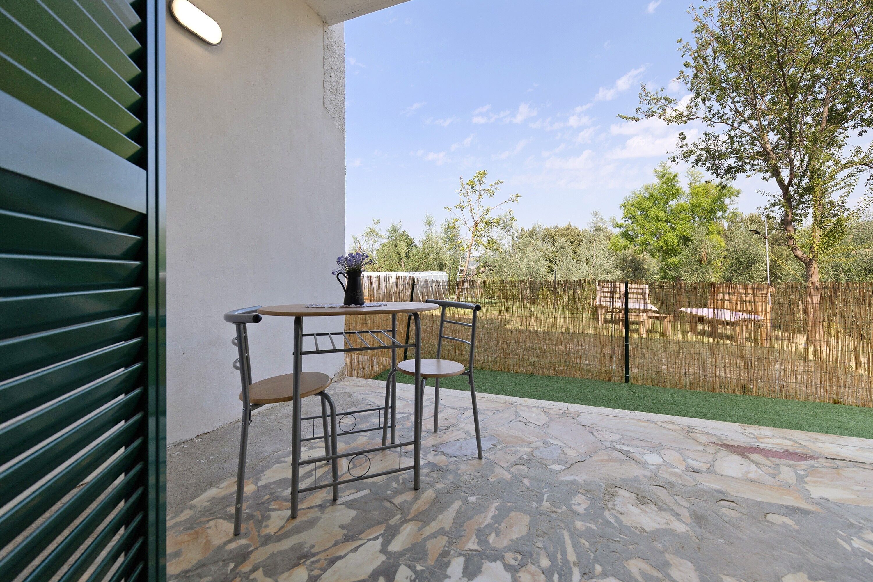 A patio with two metal chairs and a table, a small potted plant, and a view of a fenced garden with trees and a clear sky.