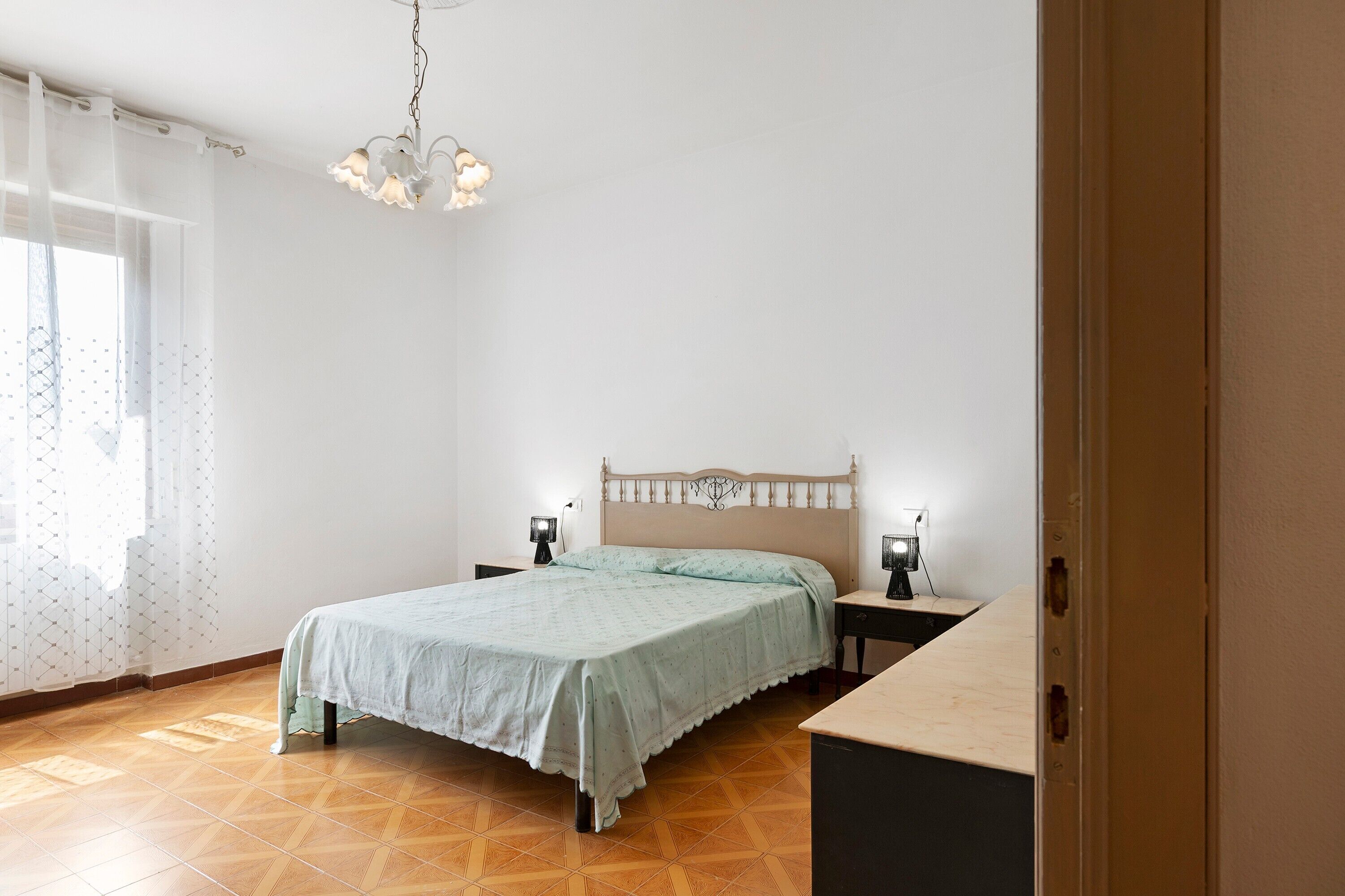 A bedroom with a bed, two bedside tables, a nightstand, a hanging light fixture, and a window with sheer curtains.