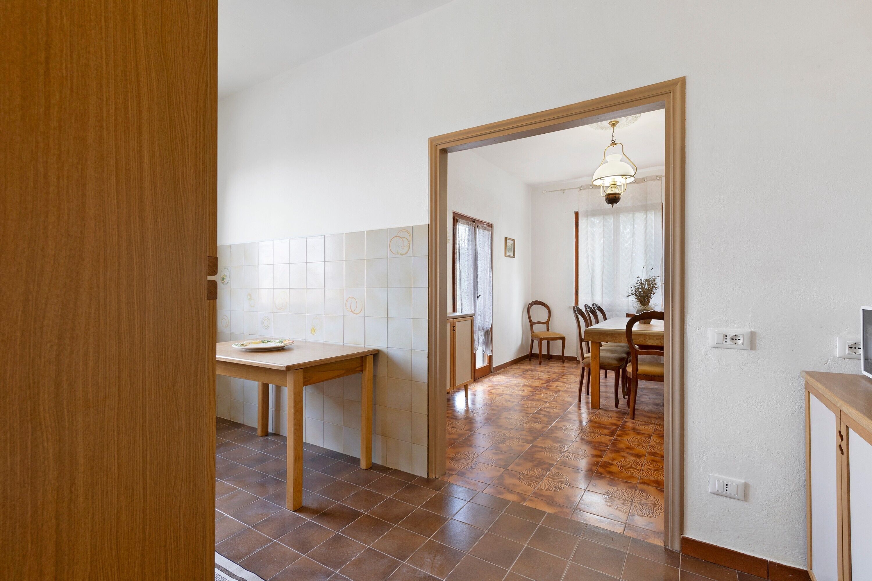 Ein Zimmer mit Holztür, einem Esstisch mit Stühlen, einer Küchenzeile mit Fliesenwand und einem Flur, der in ein weiteres Zimmer führt.