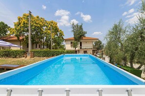 Ein rechteckiger Pool mit weißer Umrandung, umgeben von Grünflächen und einem Gebäude mit Ziegeldach.