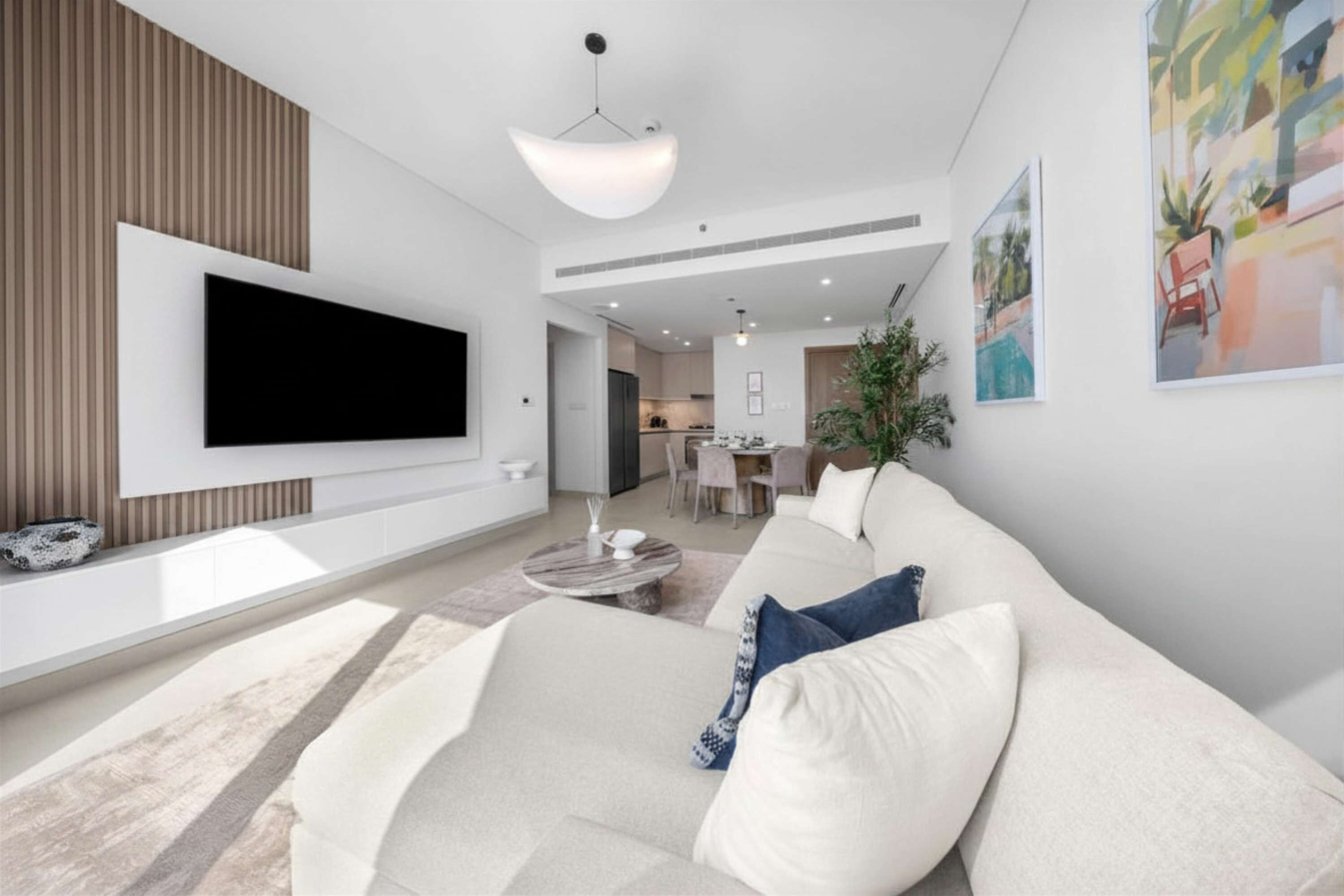 Een moderne woonkamer met een flatscreen televisie aan de muur, een hoekbank, een salontafel en een eethoek op de achtergrond.