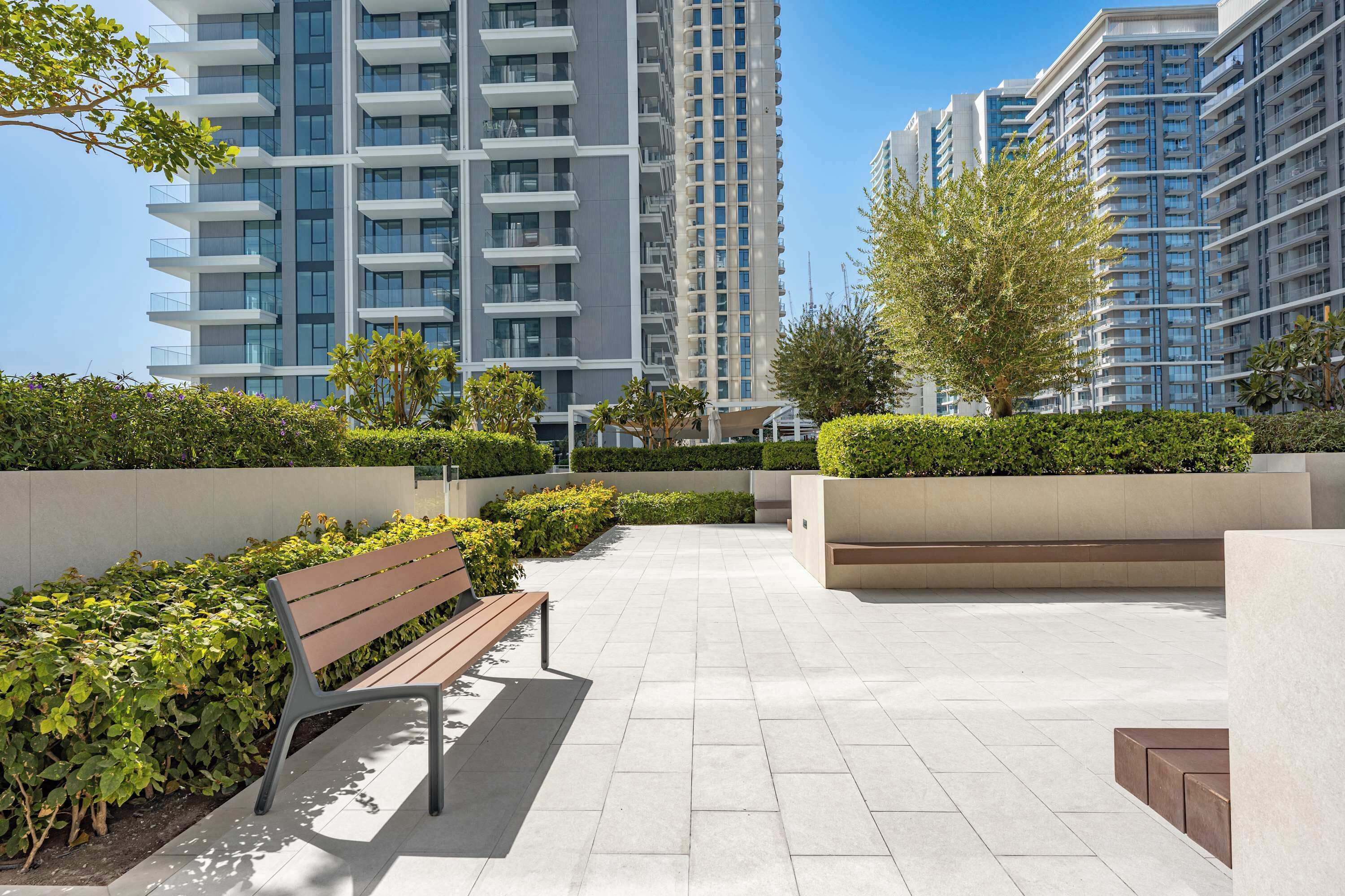 A modern outdoor area with a bench, greenery, and high-rise buildings in the background.