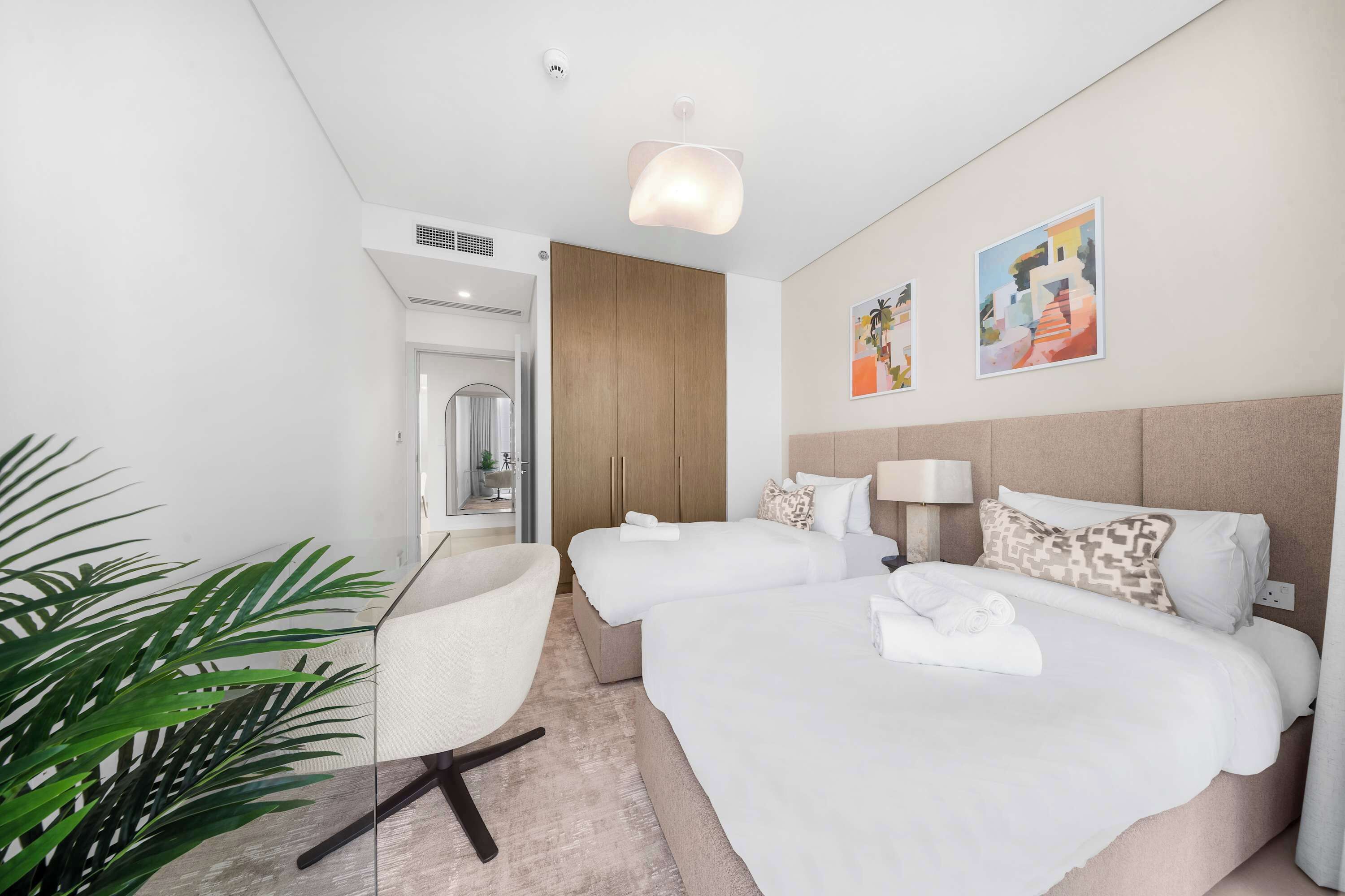 Une chambre d’hôtel avec deux lits, un bureau, une chaise, une plante et des œuvres d’art accrochées au mur.