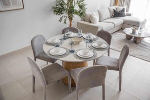 A dining area with a round table set for six, surrounded by six grey chairs, a wooden table base, and a potted plant in the background. - StayShort | Stunning Beachfront 2-Bedroom Apartment Sleeps 6 with Infinity Pool, Skyline Views & Private Beach Access (Dubai)