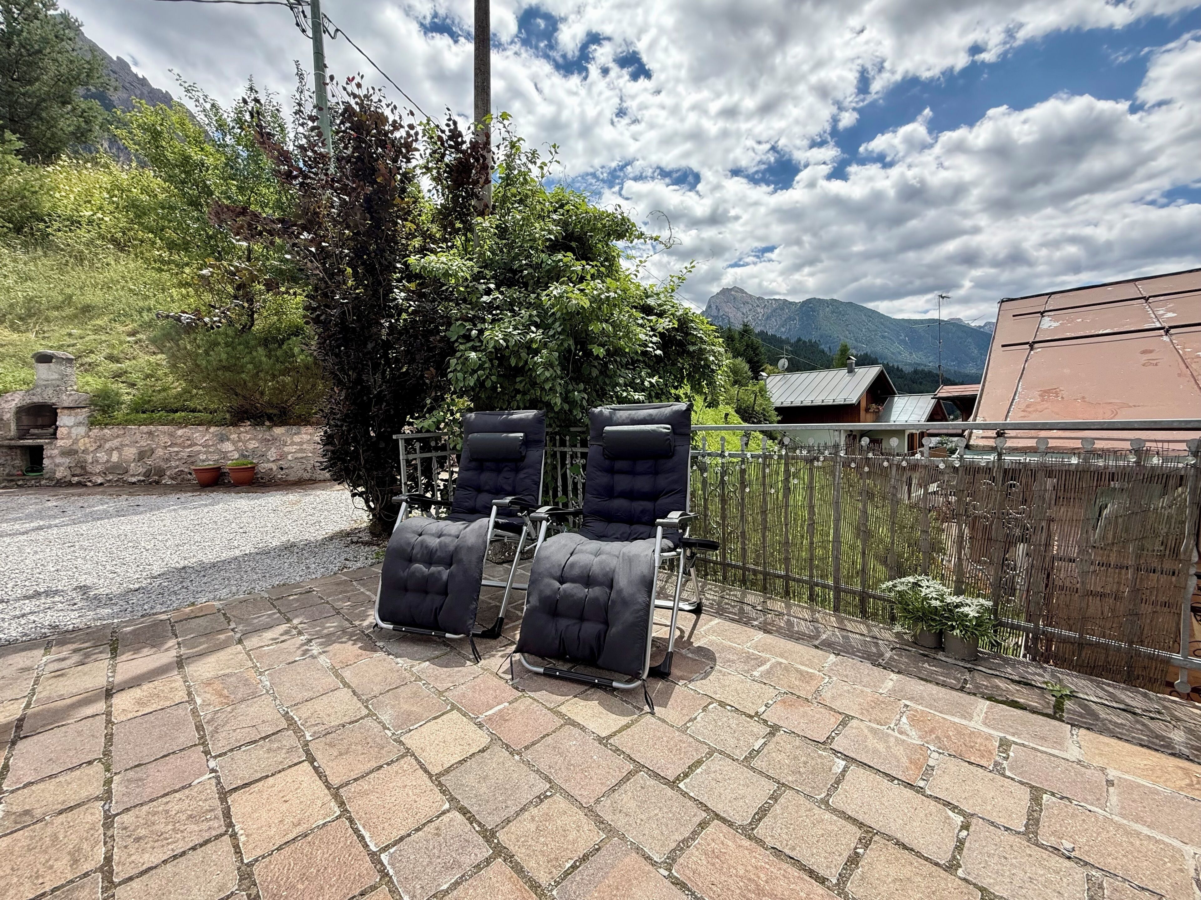 A patio with stone tiles, two black cushioned chairs, a stone wall, and a view of mountains and a house in the background.
