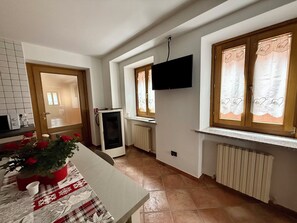 A kitchen with a table, a refrigerator, a TV mounted on the wall, and a window with curtains. - Dolomiti Cadore Home, in the heart of the Cadore Dolomites, 5 minutes from Sappada (san pietro di cadore)