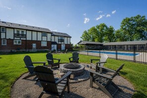 Un espace aménagé au bord de la piscine, avec des fauteuils Adirondack et un foyer, le tout entouré d’un bâtiment en briques et d’une piscine.