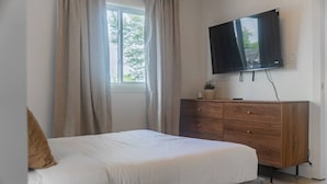 Ein Schlafzimmer mit einem Bett, einer Holzkommode, einem an der Wand montierten Fernseher und einem Fenster mit Vorhängen.