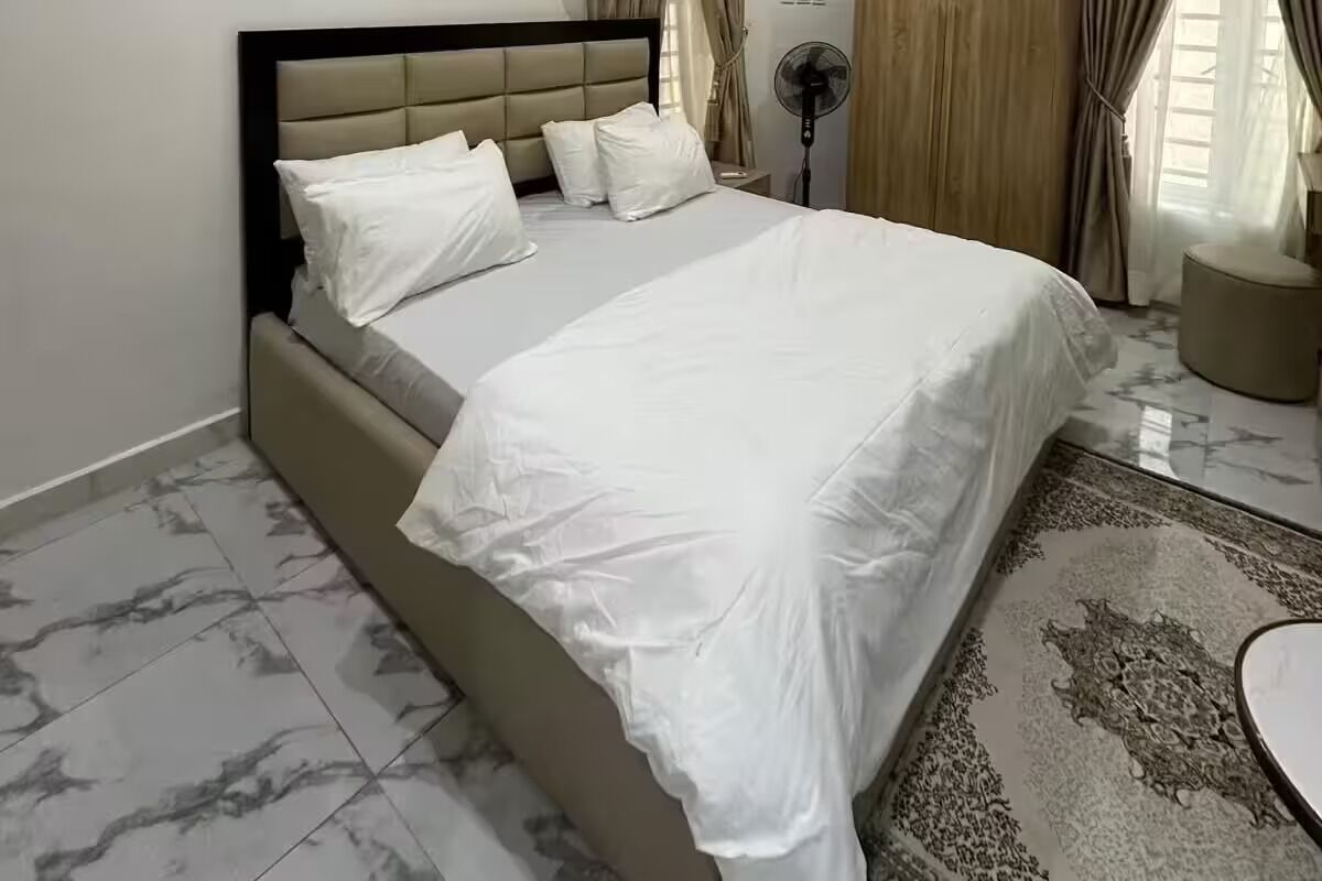 Uma cama com roupa de cama branca e cabeceira, um ventilador de chão e um tapete estampado sobre piso de azulejos.
