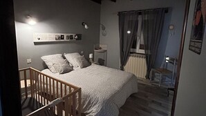 A bedroom with a bed, a nightstand, a lamp, a window with curtains, a wooden chair, and a small table. - Oustal Fraïssignol, a comfortable guesthouse (Fraisse-sur-Agout)