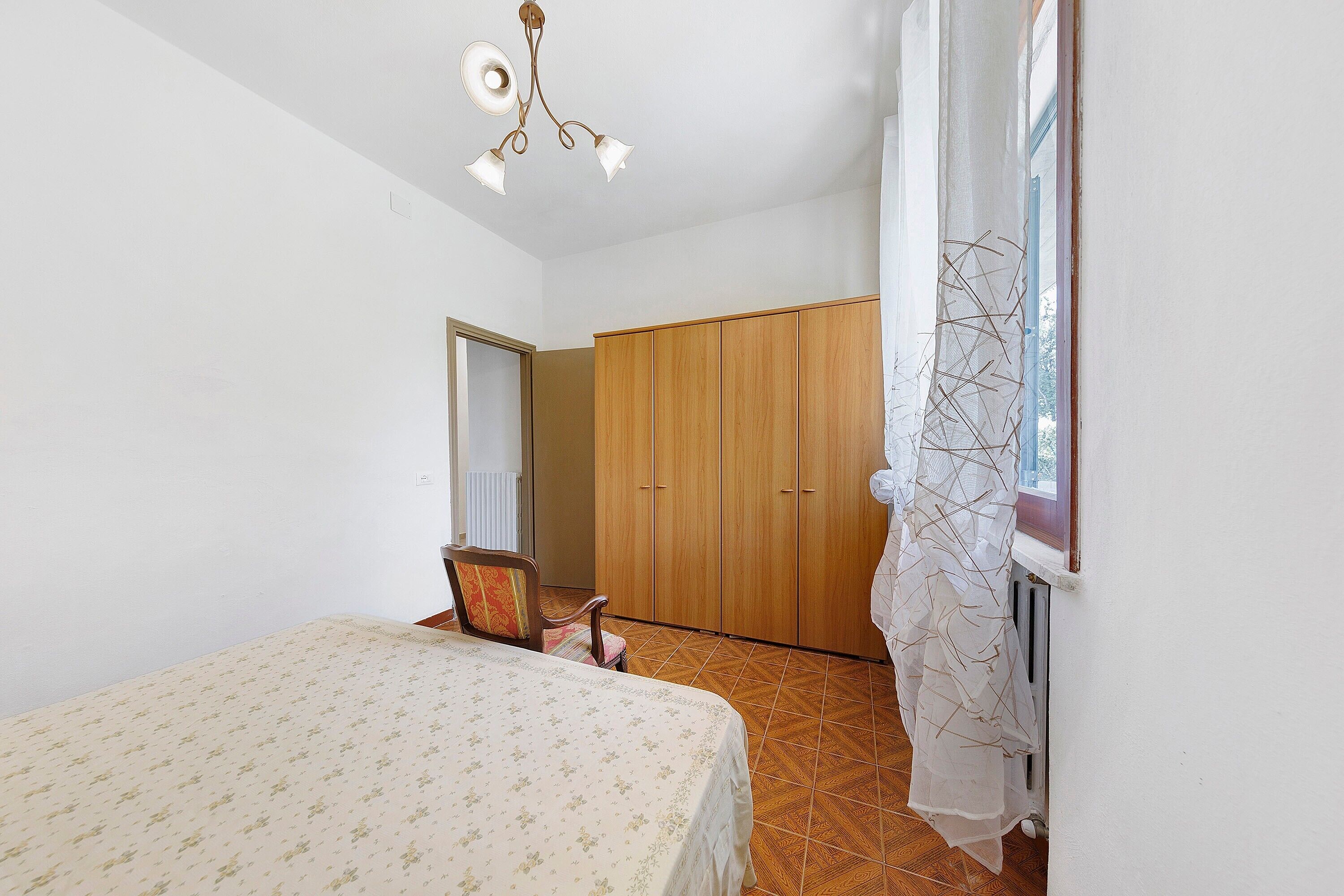 Une chambre à coucher avec un lit, une armoire en bois, une chaise et une fenêtre avec un rideau.