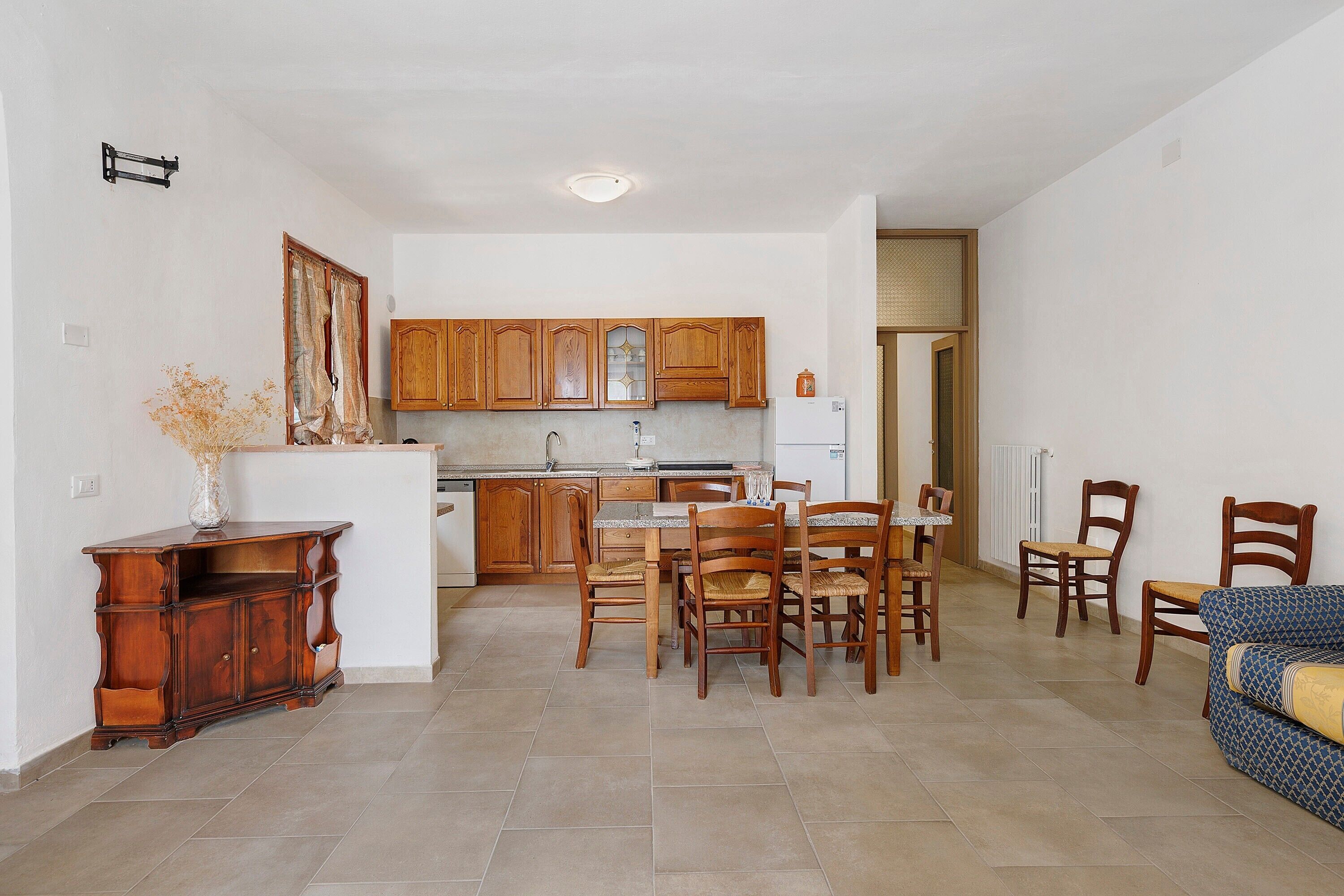 A compact kitchen with wooden cabinets, a dining area with chairs, and a small refrigerator.