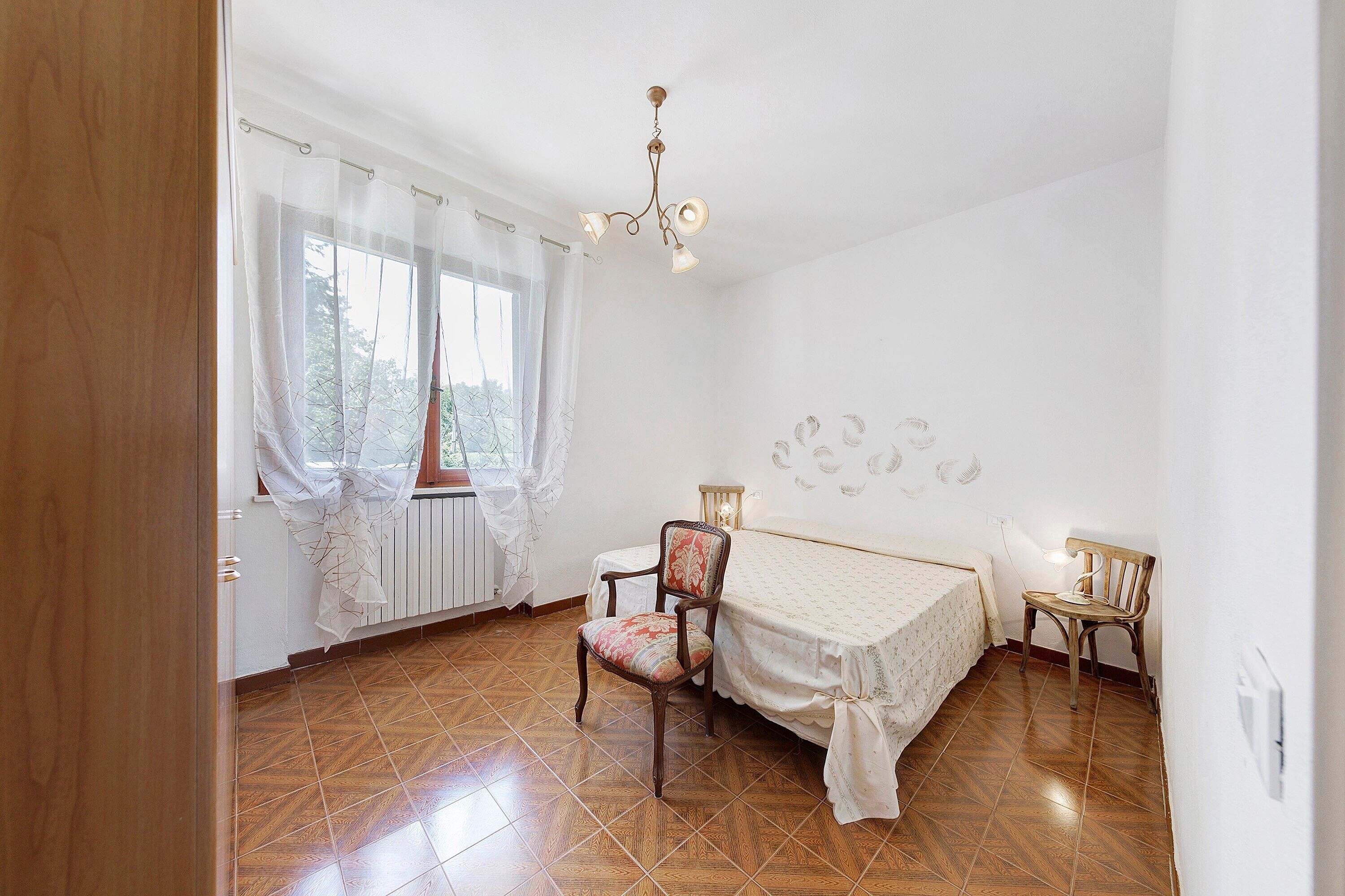Une chambre à coucher avec un lit, deux chaises, une table, un lustre et une fenêtre avec des rideaux.