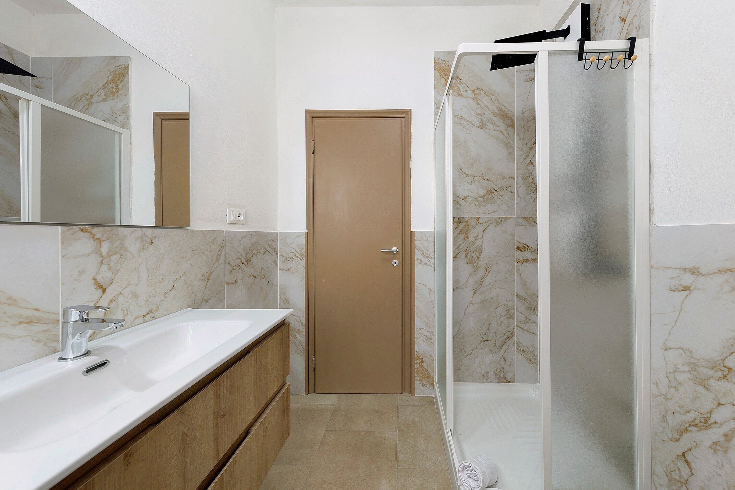 Un bagno con parete rivestita in marmo, un mobiletto in legno, un lavandino bianco e zona doccia.