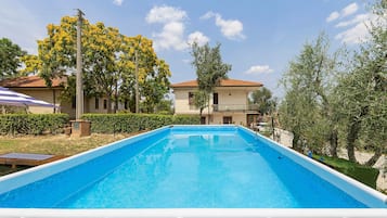 Ein rechteckiger Pool mit weißer Umrandung, umgeben von Grünflächen und einem Gebäude mit Ziegeldach.