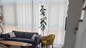 A living room with a sofa, a yellow chair, a dining table with chairs, and a large window with blinds. - Serene 3-bedroom Villa Rental in Köyceğiz, Turkey (Koycegiz)