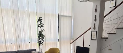 A modern living room with a sofa, a dining table with chairs, a plant, and a staircase.