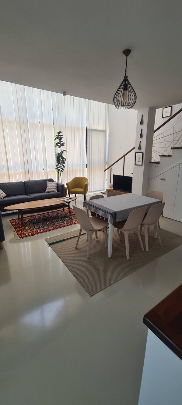 A modern living room with a sofa, a dining table with chairs, a plant, and a staircase.