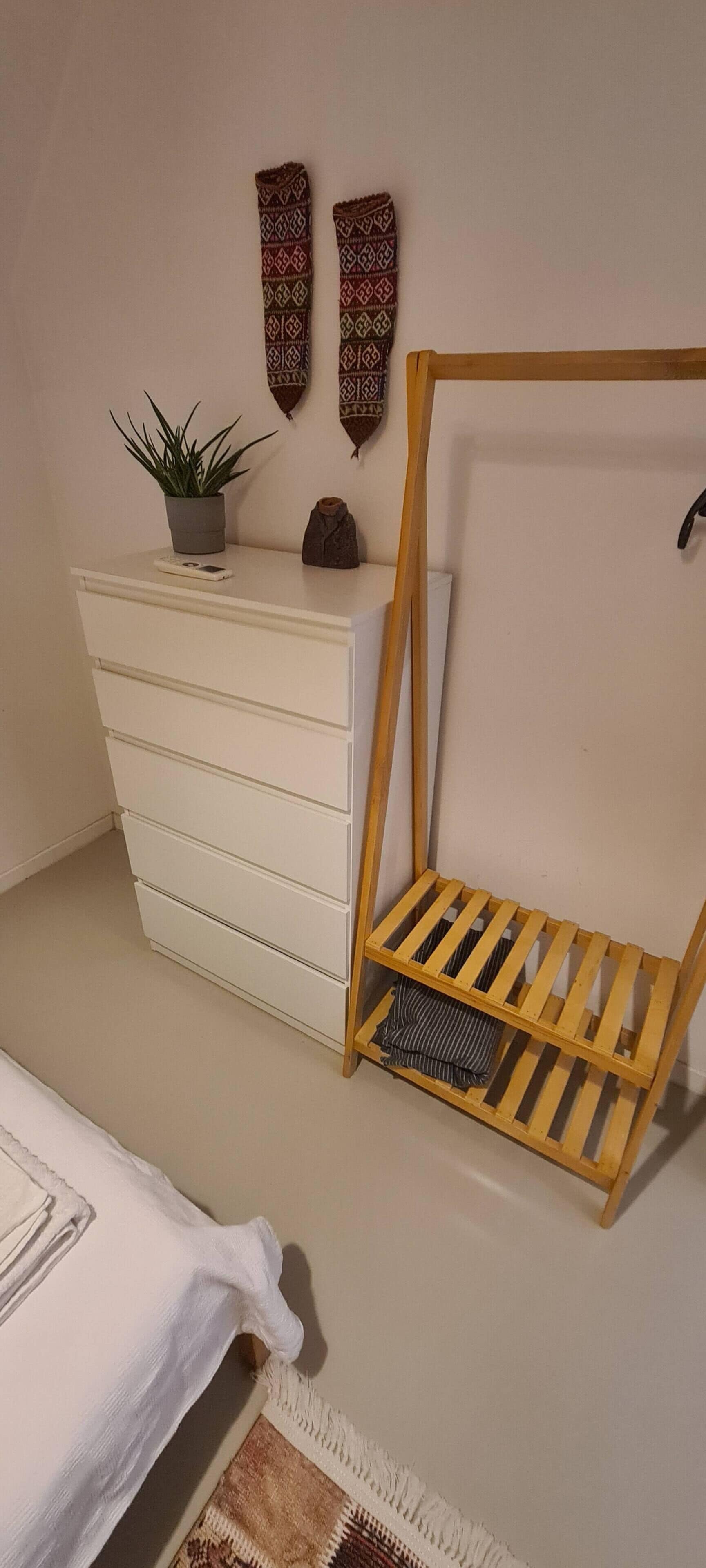 A bedroom with a white dresser, a wooden shelf, a potted plant, and a bed with a white cover.