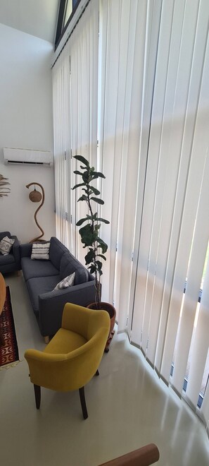 Un saló amb un sofà gris, un otomà groc, una planta en test i persianes blanques.