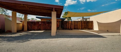 An outdoor covered patio area with a wooden roof, a concrete floor, and a sandy ground.
