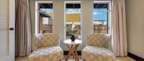 A room with two wicker chairs facing a small round table with a vase, large windows with curtains, and a view of the outside.