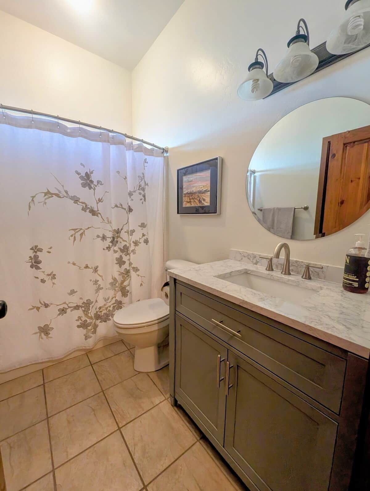 A bathroom with a toilet, a shower curtain with floral patterns, a sink with a marble countertop, a mirror, and a framed picture on the wall.