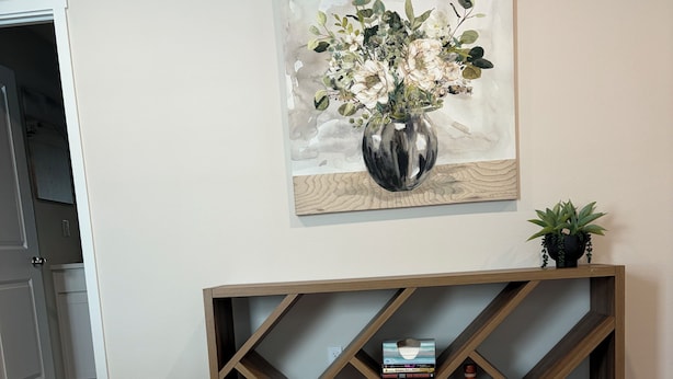 Una habitación con alfombra color beige, una consola de madera con estantes, una puerta blanca y un cuadro de flores en la pared.