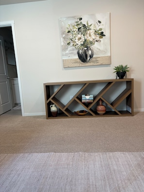 A room with a beige carpet, a wooden console table with shelves, a white door, and a painting of flowers on the wall.
