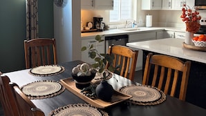 Un comedor con una mesa negra, sillas de madera y individuales tejidos. Se ve una cocina con armarios blancos y una ventana al fondo.