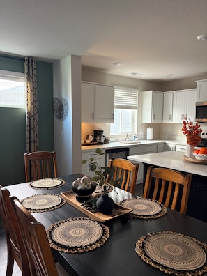 A dining area with a black table, wooden chairs, and woven placemats. A kitchen with white cabinets and a window is visible in the background. - Welcome to Your Pacific Northwest Retreat (Vancouver)