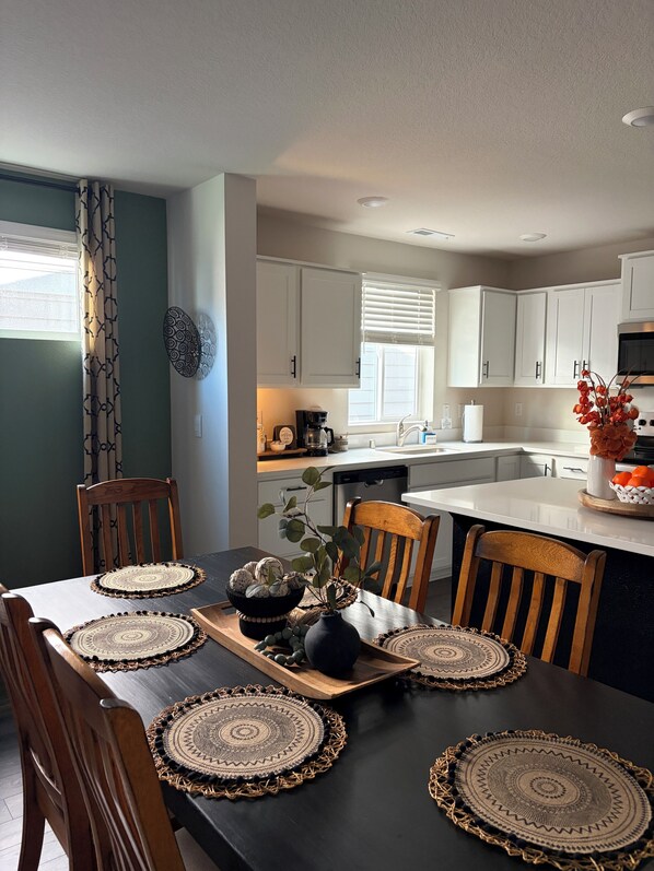 A dining area with a black table, wooden chairs, and woven placemats. A kitchen with white cabinets and a window is visible in the background.