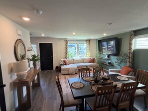 A living room with a sofa, a dining table with chairs, a TV mounted on the wall, and a door.