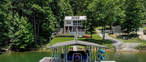 Un barco amarrado con una zona de estar cubierta, una casa con terraza y mobiliario exterior, y una orilla boscosa.