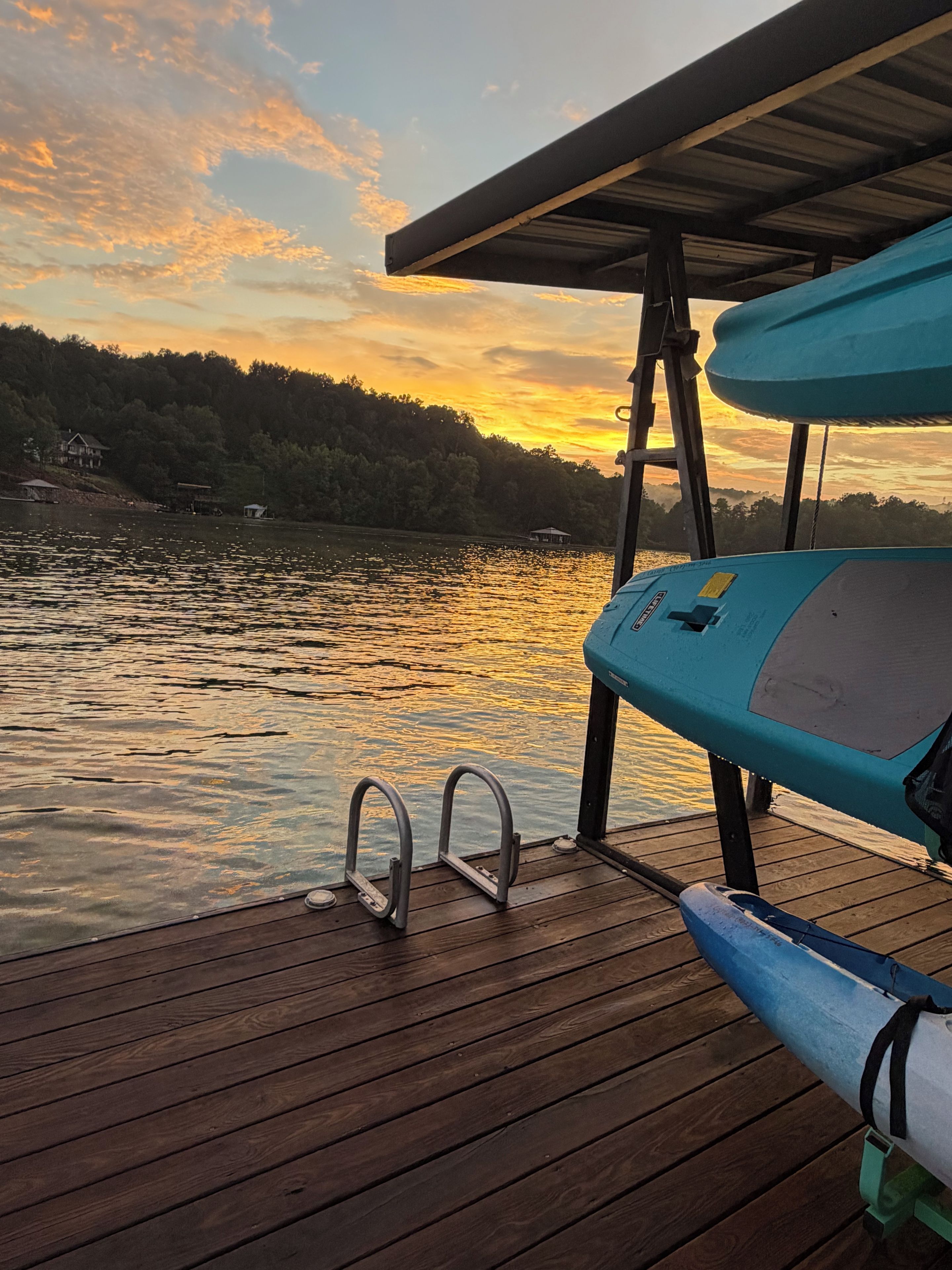 A wooden dock with metal railings, a boat, and a sunset over a forested area.