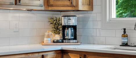 A kitchen with wooden cabinets, a coffee maker, a plant, and a window with a view of greenery.