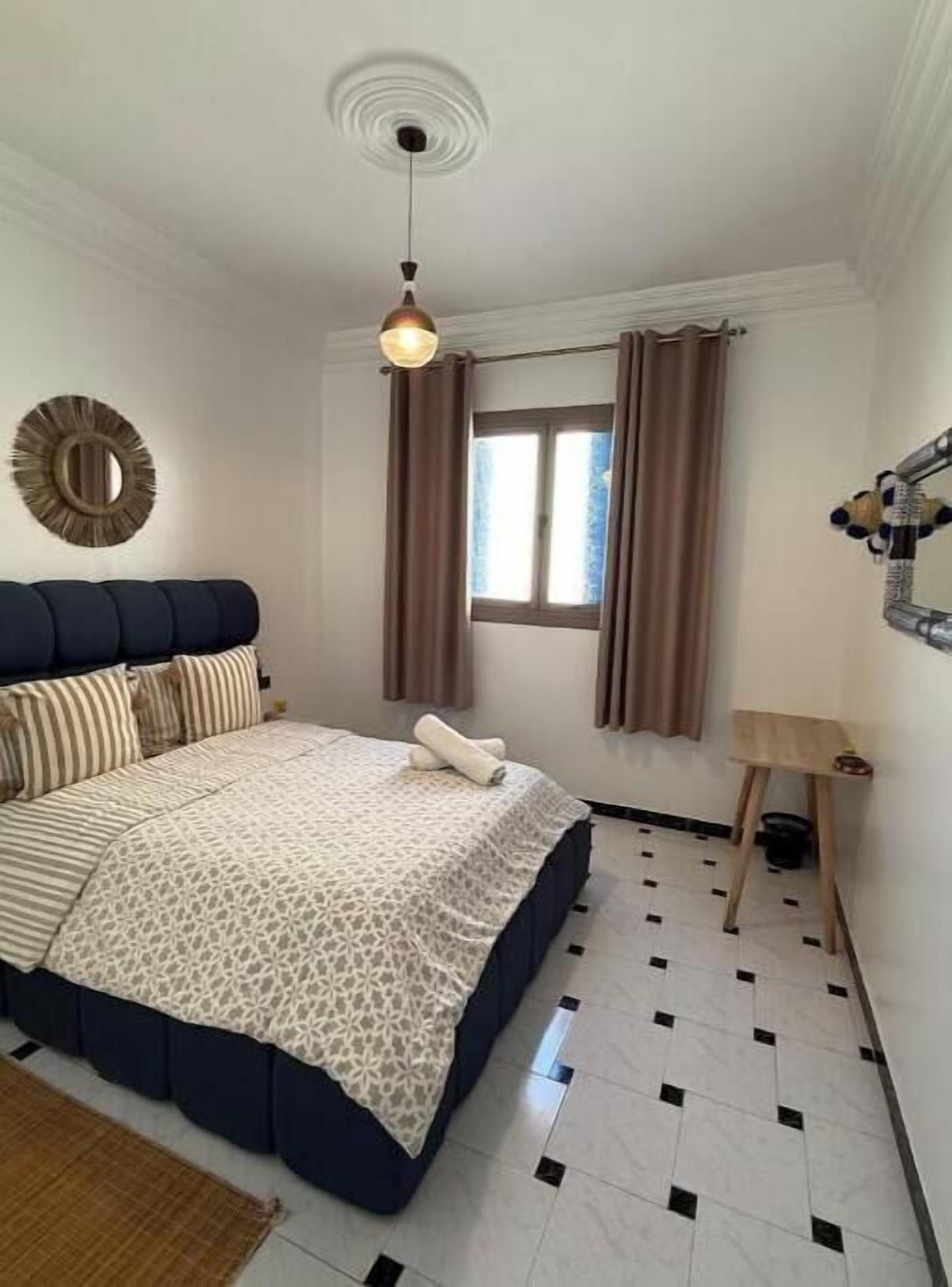 A bedroom with a bed, a hanging light fixture, a window with curtains, and a small wooden table.