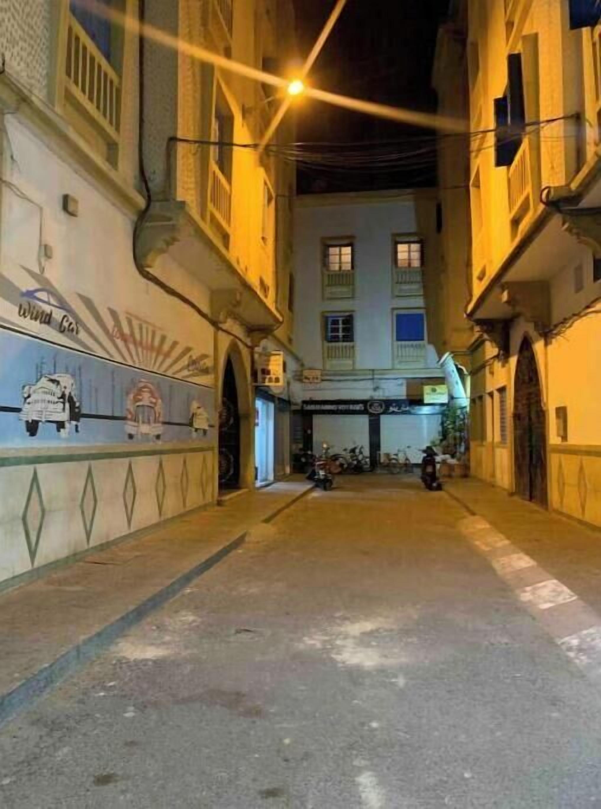 Une rue étroite, bordée de bâtiments des deux côtés, une fresque murale sur le mur de gauche et une moto garée au loin.