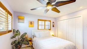 Ein Schlafzimmer mit Bett, Deckenventilator, Fensterjalousien, Wandbildern und einer Tür.