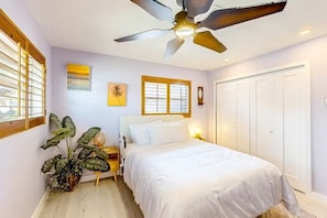Ein Schlafzimmer mit Bett, Deckenventilator, Fensterjalousien, Wandbildern und einer Tür.