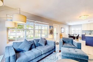 A modern living room with a sofa and armchair, large windows, and a kitchen area. - Purple Pelican Cottage | Old Florida island escape (Naples)