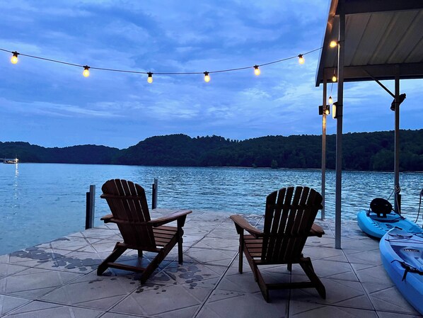 A lakeside deck with two wooden chairs, a kayak, and string lights against a backdrop of a calm lake and forested hills.
