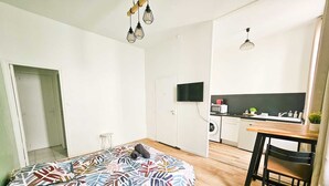 Una habitación compacta con cama, una mesita, una silla, kitchenette y televisión.