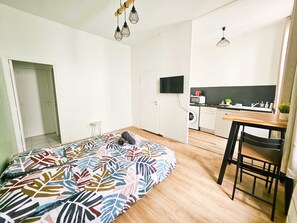Ein kleines Zimmer mit einem Bett, einem kleinen Tisch, einem Stuhl, einer Küchenzeile und einem Fernseher.