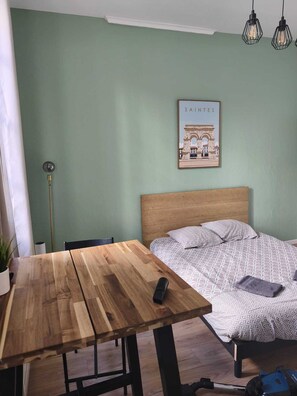 A room with a wooden table, a bed with patterned bedding, a framed picture on the wall, and a ceiling light fixture. - "Les Arches 2" City Center Studio (Saintes)