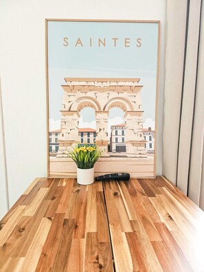 A framed artwork of the Arc de Triomphe with the word 'SAINTES' above it, placed on a wooden floor with a potted plant and a remote control beside it. - "Les Arches 2" City Center Studio (Saintes)