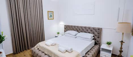Une chambre à coucher moderne avec une tête de lit capitonnée, un grand lit avec du linge de lit blanc, une table de chevet avec une lampe et une grande fenêtre avec des rideaux.