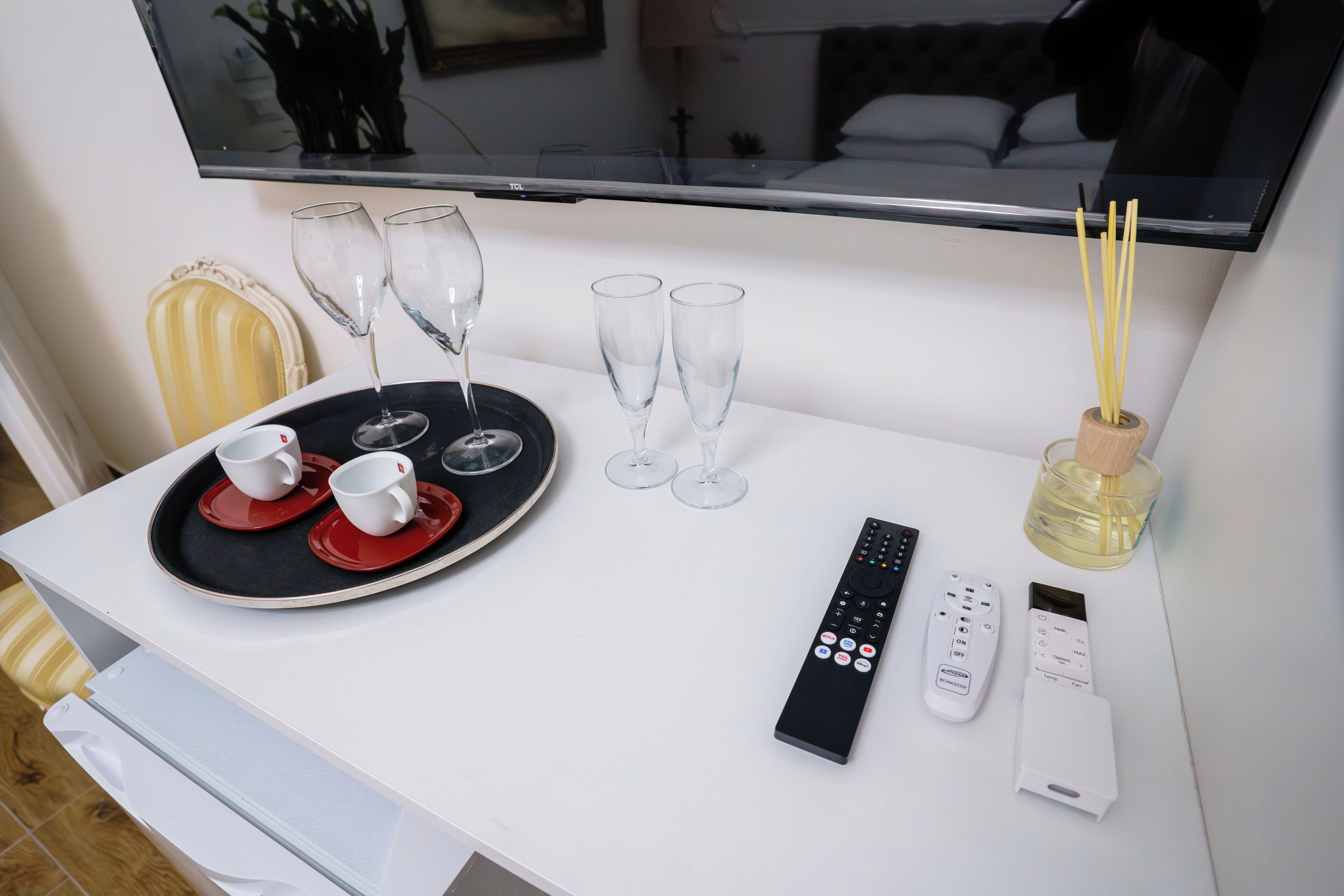 A hotel room with a white table set with two red cups, two wine glasses, and a tray. There are two remote controls and a thermostat on the table.